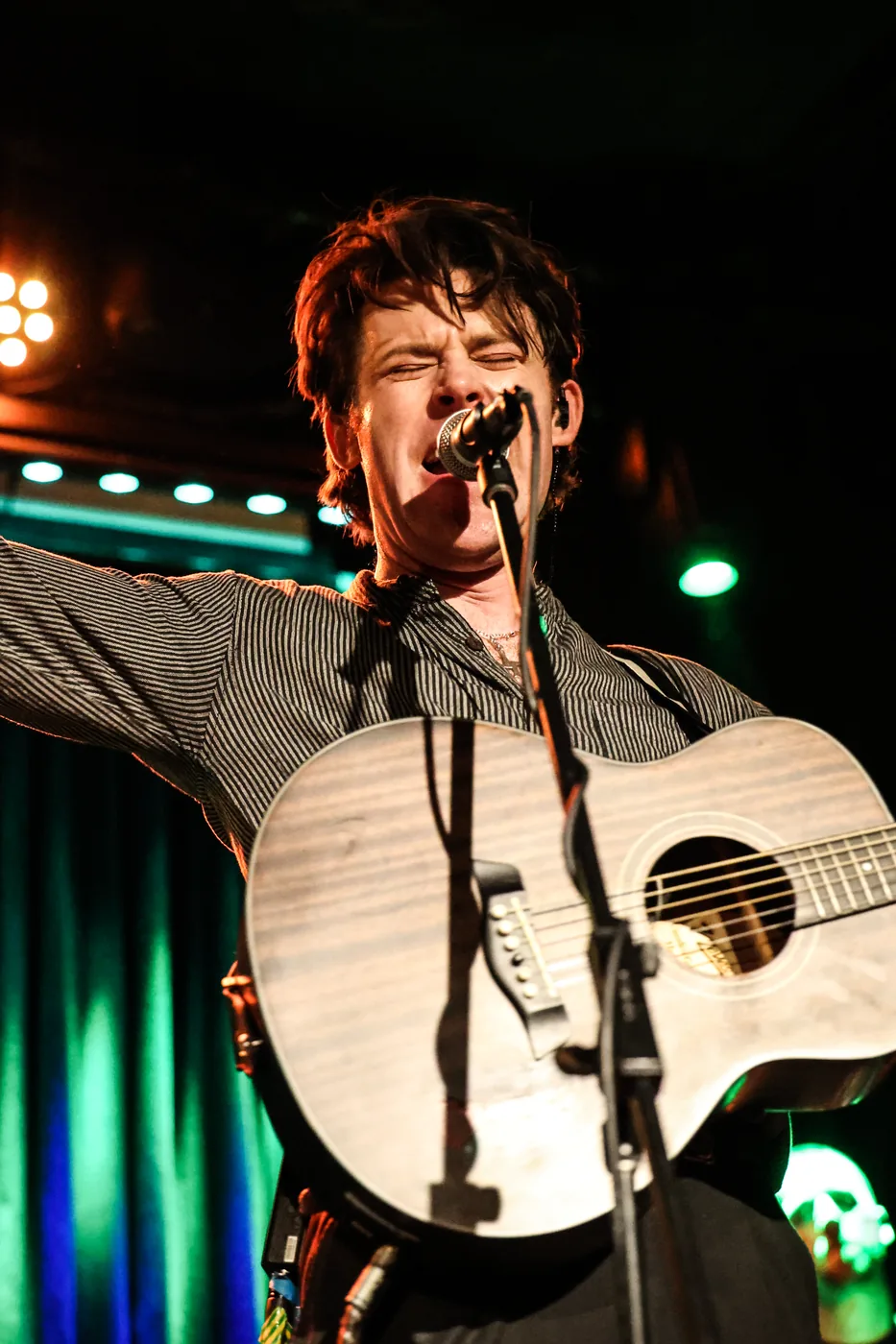 A singer at the microphone with eyes closed, holding an acoustic guitar upright, stage lights behind