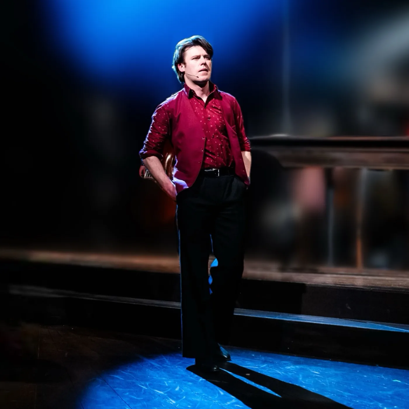 An actor in a red shirt and dark trousers standing alone in a blue spotlight on a theatre stage